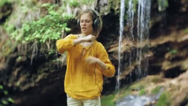スタイリッシュな美しい女性 大型モニターのヘッドフォンで音楽を聴く滝の近く — ストック動画