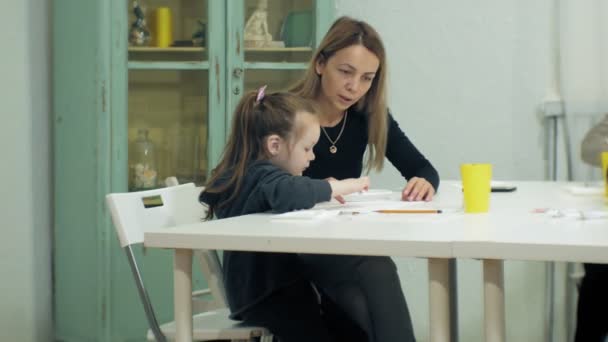 Las niñas se sientan juntas a la mesa en el aula y dibujan con los dedos y pintan. Con ellos su joven y hermosa maestra . — Vídeos de Stock
