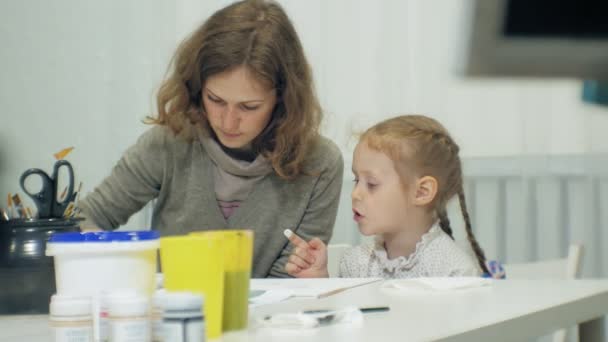 Las niñas se sientan juntas a la mesa en el aula y dibujan con los dedos y pintan. Con ellos su joven y hermosa maestra . — Vídeos de Stock