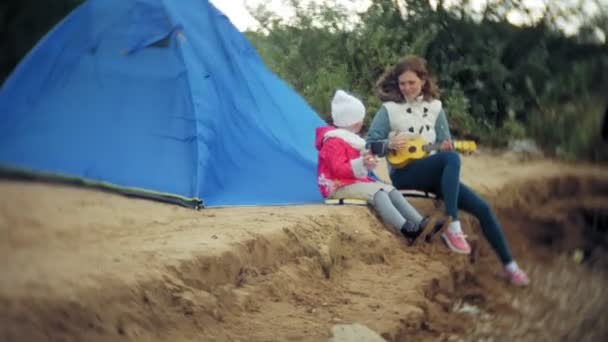 Een mooi meisje met haar familie speelt op een ukulele aan de rivieroever in de buurt van een toeristische tent — Stockvideo