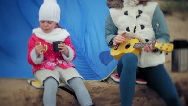 Una hermosa chica con su familia juega en un ukelele en la orilla del río cerca de una tienda de campaña turística — Vídeo de stock