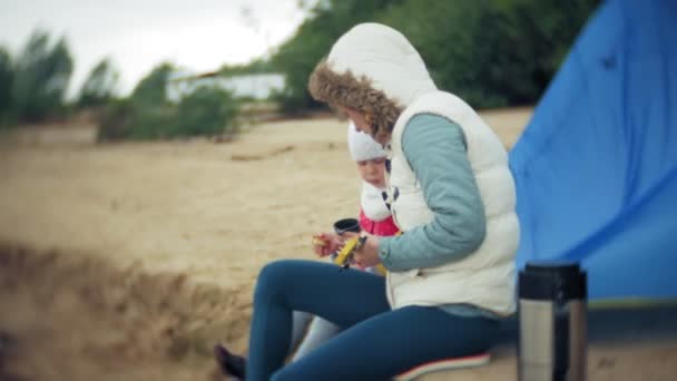 Una hermosa chica con su familia juega en un ukelele en la orilla del río cerca de una tienda de campaña turística — Vídeo de stock