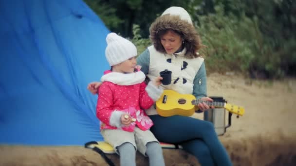 Una hermosa chica con su familia juega en un ukelele en la orilla del río cerca de una tienda de campaña turística — Vídeos de Stock