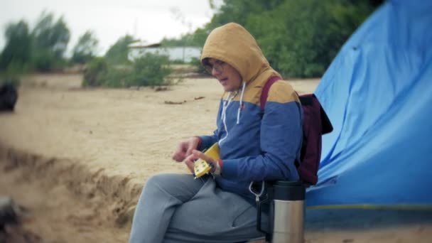 Um homem joga em um ukulele na margem do rio perto de uma tenda turística — Vídeo de Stock