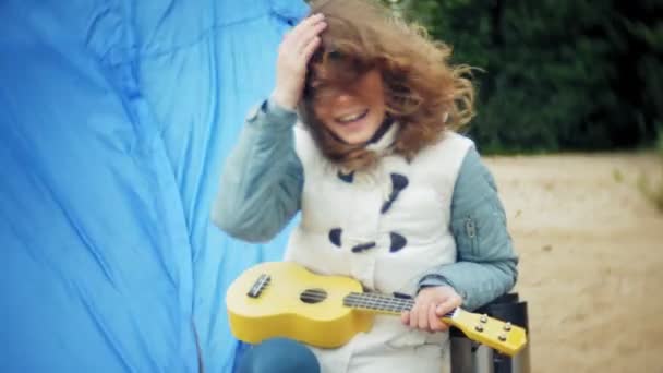 Krásná dívka s její rodinou hraje na ukulele na břehu řeky poblíž turistické stany — Stock video
