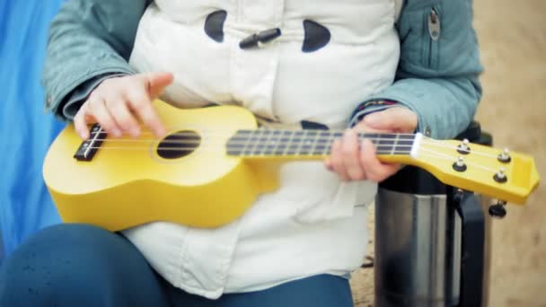 Uma menina bonita com sua família joga em um ukulele na margem do rio perto de uma tenda turística — Vídeo de Stock