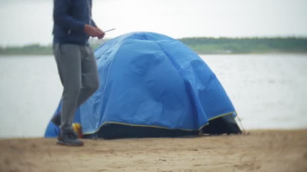 El hombre recoge una tienda de campaña turística en la orilla del río del mar — Vídeo de stock