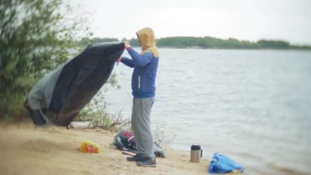 Man verzamelt een toeristische tent aan de oever van de rivier van de zee — Stockvideo