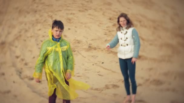 La madre juega con su hijo en un impermeable y corre por el mar en la playa — Vídeo de stock