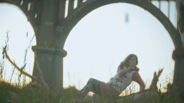 Uma jovem mulher está usando um smartphone perto de uma antiga ponte ferroviária vintage . — Vídeo de Stock