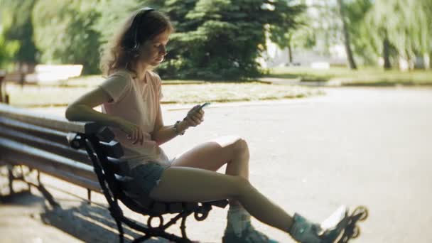 Mujer patinando y escuchando música en los auriculares en el teléfono en el parque — Vídeos de Stock