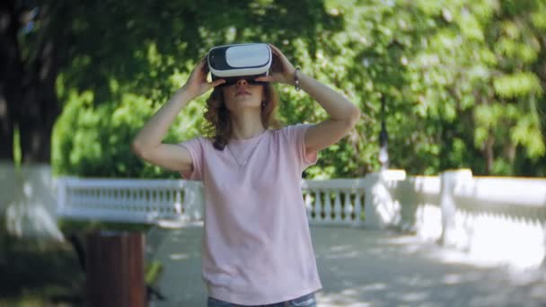 Mujer joven utiliza la pantalla montada en la cabeza en el parque. Jugar a juego con casco VR para teléfonos inteligentes. Feliz. — Vídeos de Stock