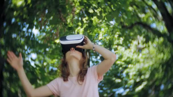 Young woman uses head-mounted display in the park. Playing game using VR-helmet for smart phones. Happy — Stock Video