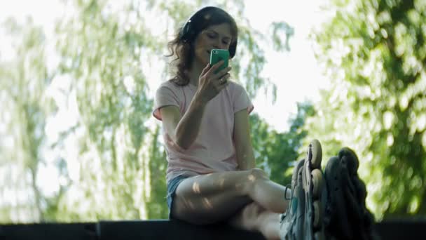 Жінка-ролики та прослуховування музики на навушниках на телефоні в парку — стокове відео