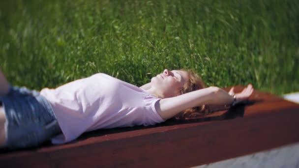 Красива дівчина лежить на лавці парку і мріє. Насолоджуйтесь природою Завершення відтворення з повільним рухом — стокове відео