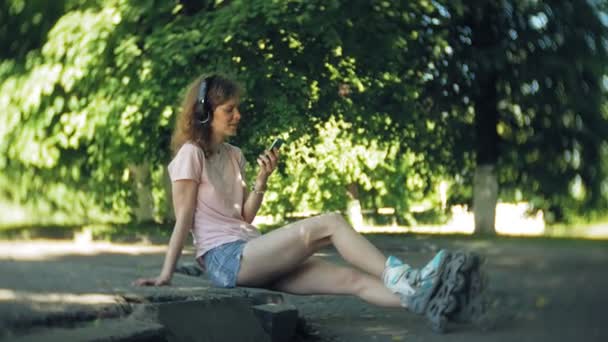 Woman rollerblading and listening to music on headphones on the phone in the park — Stock Video