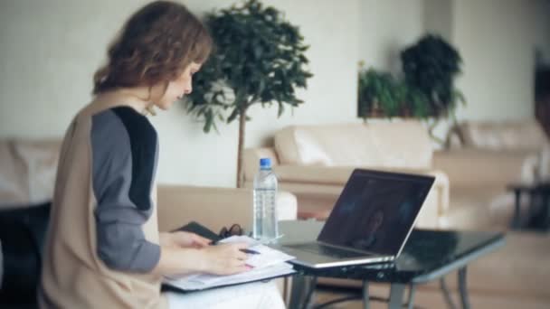 Joven mujer de negocios hermosa sentada en el sofá en la mesa con la tableta y mirando a través de papel — Vídeos de Stock