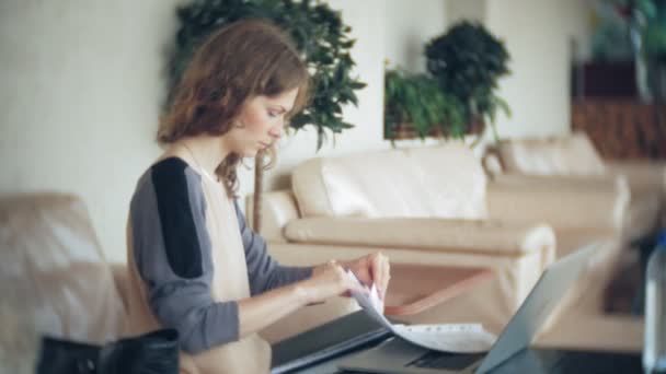 Joven mujer de negocios hermosa sentada en el sofá en la mesa con la tableta y mirando a través de papel — Vídeo de stock