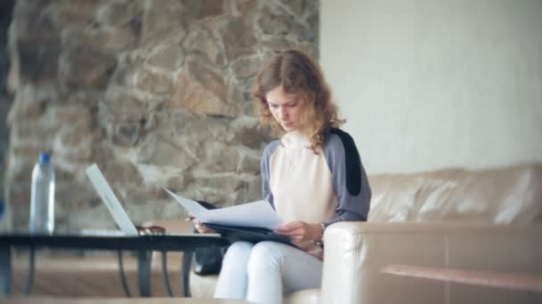 Young beautiful business woman sitting on sofa at table with tablet and looking through paper — Stock Video