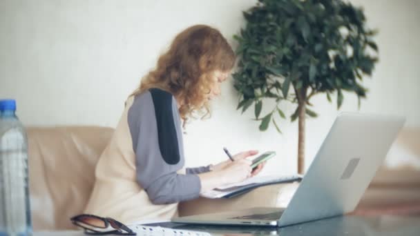 Jovem mulher de negócios bonita sentada no sofá à mesa com tablet e usando smartphone — Vídeo de Stock