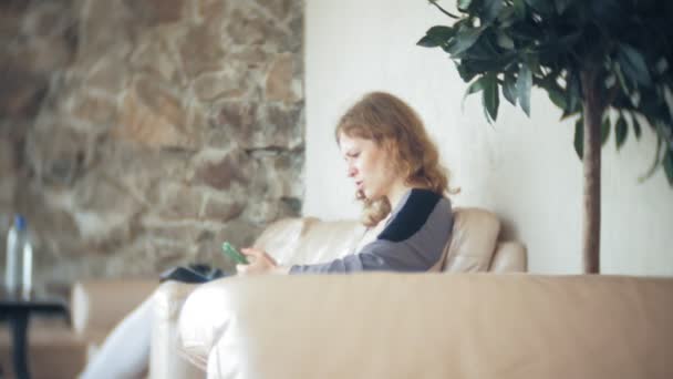 Jovem mulher de negócios bonita sentada no sofá à mesa com tablet e usando smartphone — Vídeo de Stock