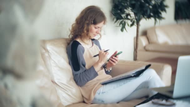 Jeune belle femme d'affaires assise sur un canapé à table avec tablette et utilisant un smartphone — Video