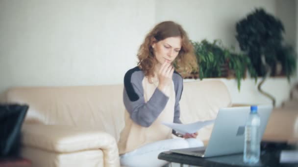 Joven mujer de negocios hermosa sentada en el sofá en la mesa con la tableta y mirando a través de papel — Vídeo de stock