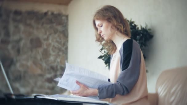 Joven mujer de negocios hermosa sentada en el sofá en la mesa con la tableta y mirando a través de papel — Vídeo de stock