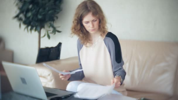 Joven mujer de negocios hermosa sentada en el sofá en la mesa con la tableta y mirando a través de papel — Vídeos de Stock