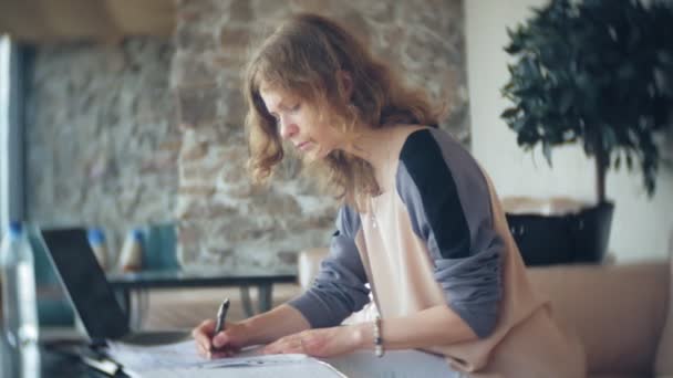 Молода красива бізнес-леді сидить на дивані за столом з планшетом і дивиться через папір — стокове відео