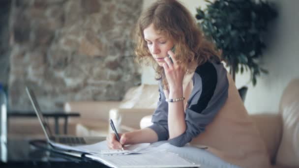 Joven mujer de negocios hermosa sentada en el sofá en la mesa con la tableta y mirando a través de papel — Vídeos de Stock