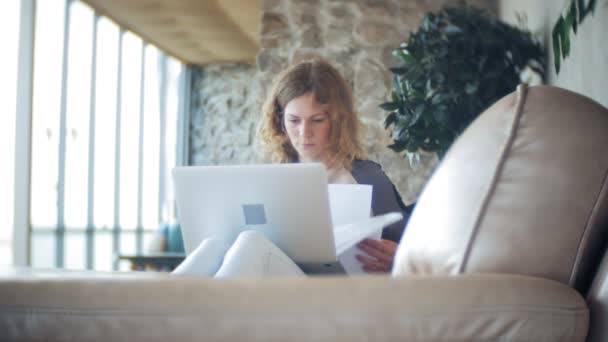 Молода красива бізнес-леді сидить на дивані за столом з планшетом і дивиться через папір — стокове відео