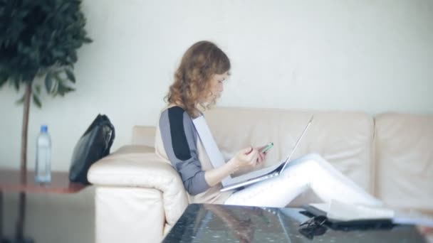 Jovem mulher de negócios bonita sentada no sofá à mesa com tablet e olhando através do papel — Vídeo de Stock