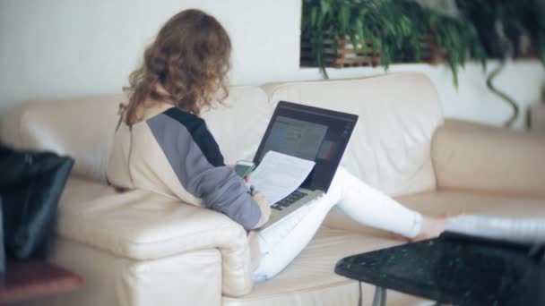 Joven mujer de negocios hermosa sentada en el sofá en la mesa con la tableta y mirando a través de papel — Vídeos de Stock