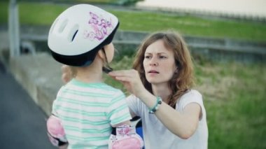 Anne kızını bir kask ve parkta paten için koruyucu giysiler giymek için yardımcı olur. Kadın koruyucu dizine koymak ve dirsek pedleri kız yardımcı olur. Parkta aktif aile dinlenme.