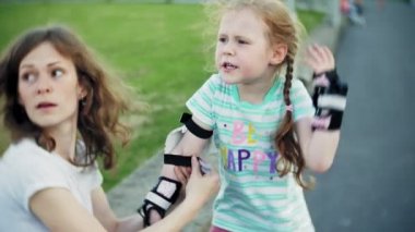 Anne kızını bir kask ve parkta paten için koruyucu giysiler giymek için yardımcı olur. Kadın koruyucu dizine koymak ve dirsek pedleri kız yardımcı olur. Parkta aktif aile dinlenme.