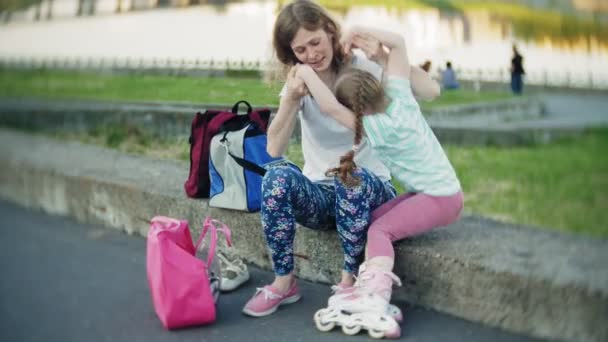 Madre ayuda a su hija a usar un casco y equipo de protección, para patinar en el parque. La mujer ayuda a la niña a ponerse protectores de rodilla y coderas. Descanso familiar activo en el parque . — Vídeos de Stock