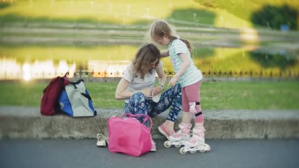 Moeder helpt dochter te dragen van een helm en beschermende kleding, voor rolschaatsen in het park. Vrouw helpt meisje zetten beschermende knie en elleboog pads. Actieve familie rust in het park. — Stockvideo