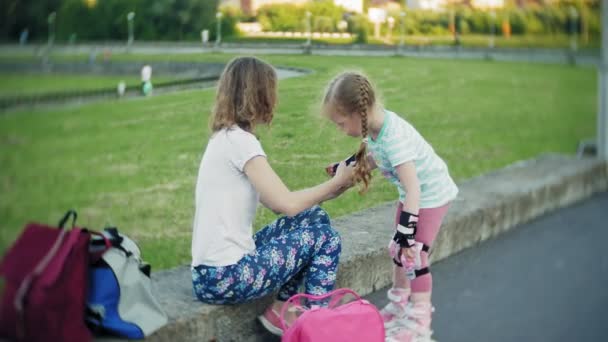 ヘルメットと公園にローラー スケート用の防護服を着用する彼の娘を母に役立ちます。女性の少女保護の膝の上に置くし、パッドを肘に役立ちます。公園でアクティブな家族の残り. — ストック動画