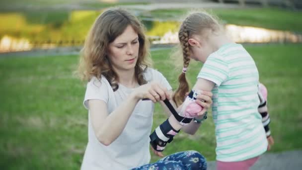 Matka pomaga jego córka nosić kask i ochraniacze/kaski, na rolkach w parku. Kobieta pomaga dziewczyna na ochronne kolana i łokcie. Aktywny wypoczynek dla rodzin z dziećmi w parku. — Wideo stockowe