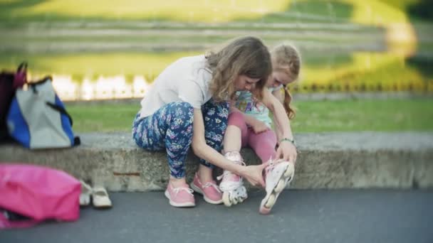 Matka pomůže jeho dcera nosit přilbu a chrániče pro bruslaře v parku. Žena pomáhá dívka na ochranné kolenní a loketní chrániče. Rodinná relaxace v parku. — Stock video