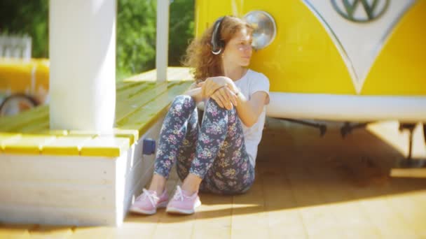 黄色のヴァゴン車音楽を聴くヘッドフォン スマート フォンを使用して -、リラックスを楽しむ、技術の旅行の概念ごと屋外に若い女性 — ストック動画
