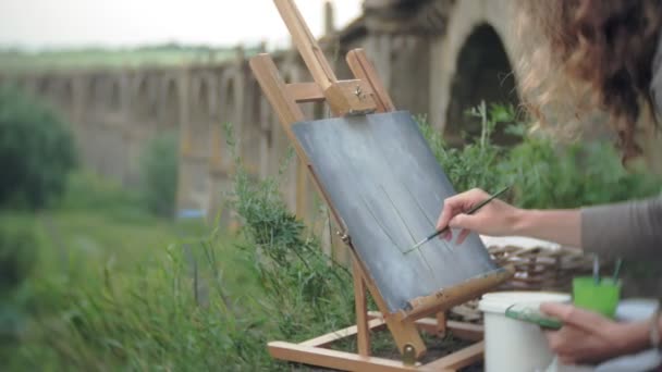 Mujer joven se basa en las pinturas de caballete y cepillo viejo puente vintage — Vídeos de Stock