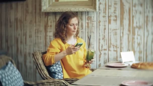 Молода жінка п'є коктейль у кафе-барі і користується телефоном — стокове відео