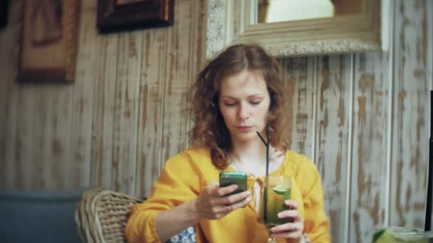 Eine junge Frau trinkt einen Cocktail in einer Cafébar und benutzt ein Telefon — Stockvideo