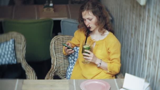 Mladá žena pije koktejl v café baru a používá telefon — Stock video
