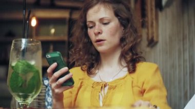 Genç bir kadın bir kafe barda bir kokteyl içiyor ve bir telefon kullanır