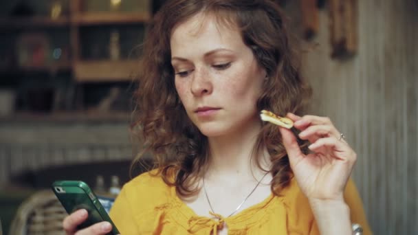 Uma jovem come uma torta em um café bar e usa um telefone — Vídeo de Stock