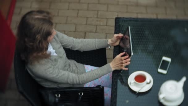 Giovane donna utilizza un tablet e telefono, beve il tè in un bar — Video Stock