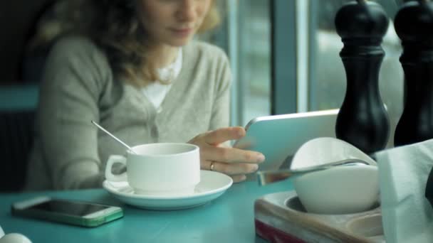 Νεαρή γυναίκα χρησιμοποιεί ένα tablet και τηλέφωνο, πίνει τσάι σε ένα καφέ-μπαρ — Αρχείο Βίντεο